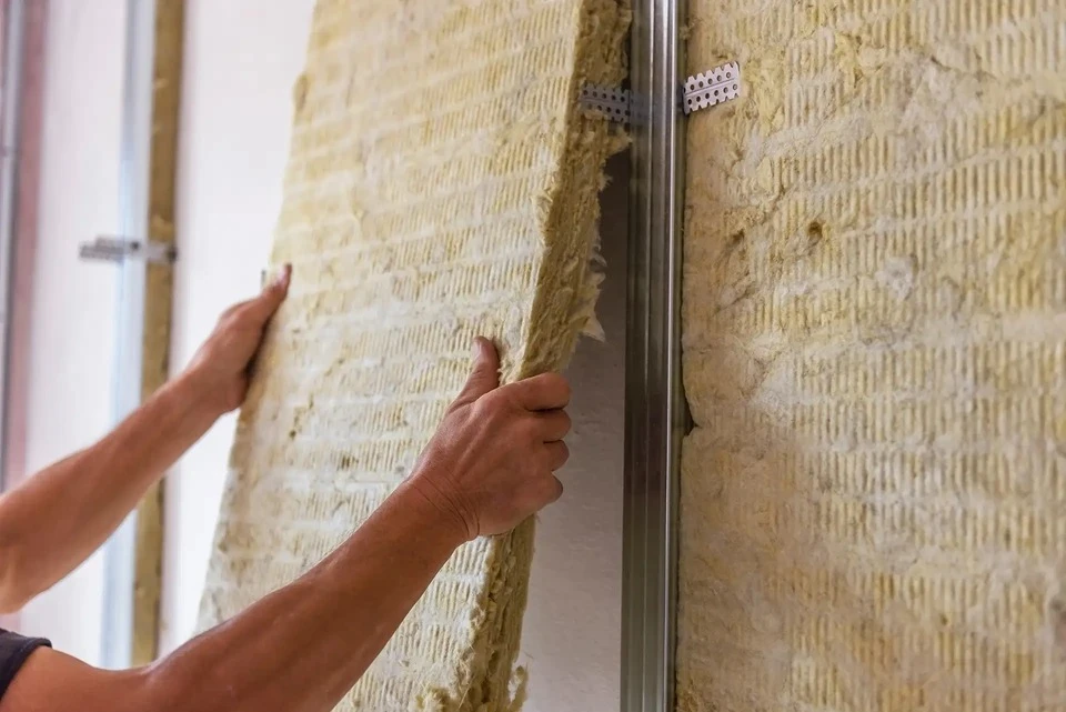 Deux ou trois artisans/ouvriers du bâtiment en tenue professionnelle travaillant ensemble sur un chantier intérieur de pose de plaques de plâtre. L'un d'eux est en train de fixer ou de poser une plaque de plâtre sur une cloison, tandis qu'un autre l'assiste ou prépare le chantier. La scène doit montrer le travail méthodique et soigné, l'interaction collaborative entre les membres de l'équipe, et metttre en évidence le professionnalisme du savoir-faire en plâtrerie. Arrière-plan : intérieur d'un bâtiment en cours de rénovation/construction.